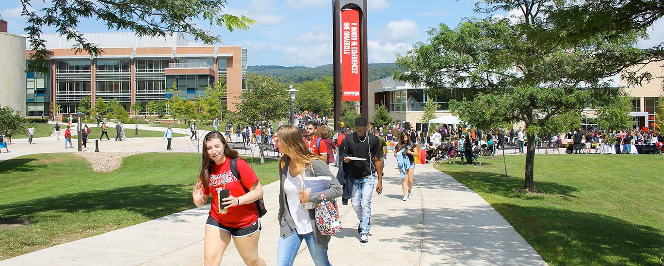 A large group of Frostburg State students walk to class near the campus clocktower on a warm, sunny day
